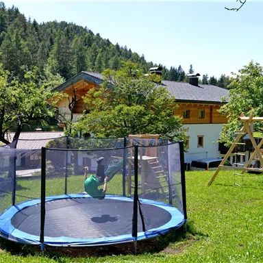 Ein Trampolin auf einer grünen Wiese, umgeben von Bäumen und einem Spielplatz. Im Hintergrund ist ein großes, helles Haus zu sehen.