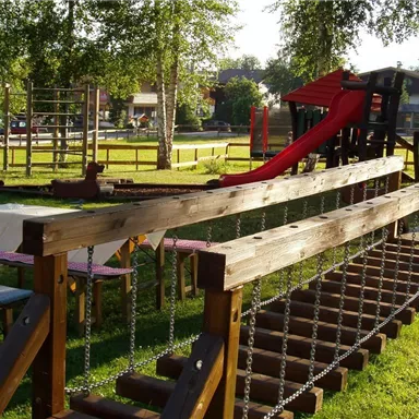 Ein Spielplatz mit einer Rutsche und einem Klettergerüst in einer grünen Parklandschaft. Tische stehen bereit für Besucher, die die Natur genießen möchten.