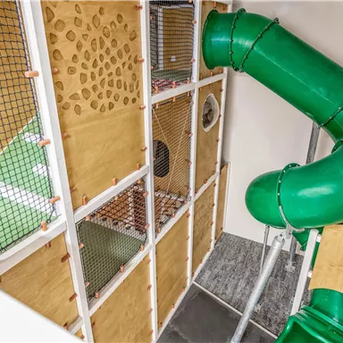A play area with a green slide tunnel and various climbing elements. The walls are made of wood with net and hole patterns.
