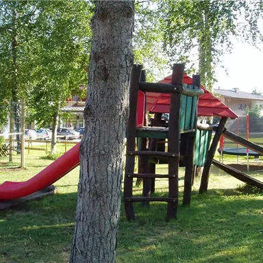 Ein Spielplatz mit einer Rutsche und einem Spielturm aus Holz. Grünes Gras und Bäume umgeben die Spielgeräte.