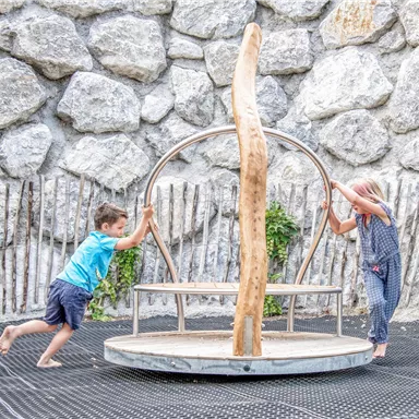 Two children are playing on a playground structure outdoors. In the background, a stone wall can be seen.