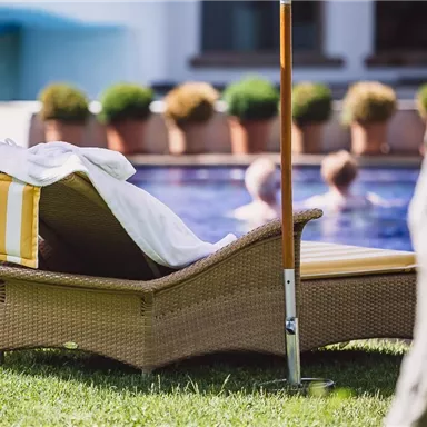 A deck chair with yellow-and-white striped towels lies on the lawn. In the background, swimmers are visible in the pool.