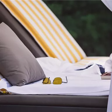 A comfortable lounge chair with striped cushions and a white blanket. On the table lies a pair of sunglasses and a book.