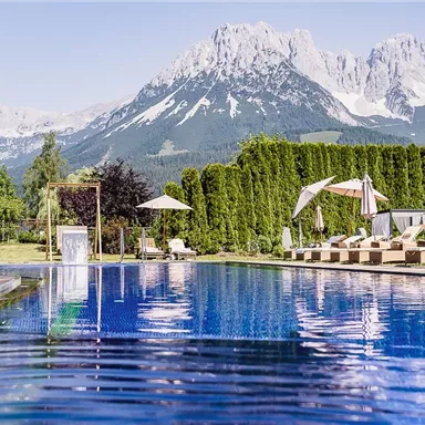 A beautiful pool with clear water and loungers surrounded by green trees. In the background, impressive mountains and a blue sky can be seen.