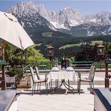 A beautiful outdoor area with a table and chairs, surrounded by blooming plants. In the background, impressive mountains can be seen.