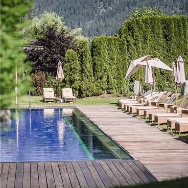 A beautiful pool with a wooden deck and sun loungers. Behind the pool, green hedges and mountains can be seen.