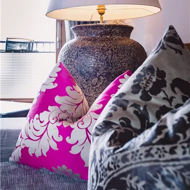 A cozy living room with colorful cushions and a decorative lamp. In the background, there is a stylish vase.