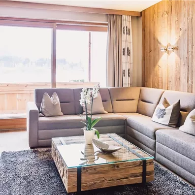 A cozy living room with a modern couch and a wooden table. Large windows offer a view of the nature outside.