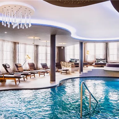 An elegant wellness area with a large swimming pool and comfortable loungers. The room is brightly lit and furnished with stylish design elements.