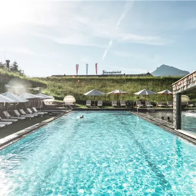 A beautiful pool with loungers and umbrellas surrounded by green meadows. In the background, mountains and a clear sky can be seen.