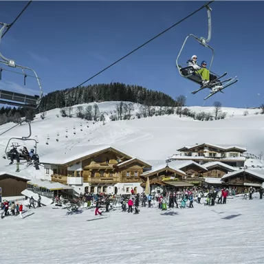 Ein Skigebiet mit schneebedeckten Bergen und einer Skilift-Anlage. Im Vordergrund sind zahlreiche Skifahrer und eine gemütliche Berghütte zu sehen.