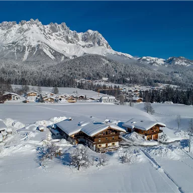 Eine winterliche Landschaft mit schneebedeckten Hütten und majestätischen Bergen im Hintergrund. Der klare blaue Himmel hebt die ruhige Atmosphäre der Szene hervor.