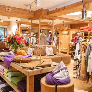 A modern clothing store with wooden shelves and colorful garments. Shoes and decorative flower arrangements are visible on a table.
