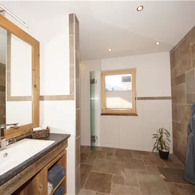 Ein modernes Badezimmer mit einem großen Spiegel und einer eleganten Steinfliese. Das Licht fällt durch ein Fenster und es gibt einen dekorativen Pflanzenarrangement.