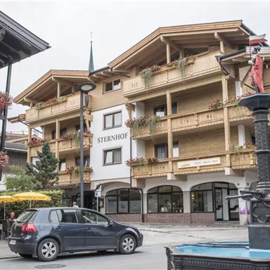 Ein charmantes Gebäude mit dem Namen "Sternhof", das traditionell mit Balkonen und Blumen geschmückt ist. Im Vordergrund sieht man einen Brunnen und eine Straße mit wenigen Autos.