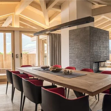 Ein moderner Essbereich mit einem großen Holztisch und roten Stühlen. Die großen Fenster bieten einen Blick auf die umliegende Landschaft.