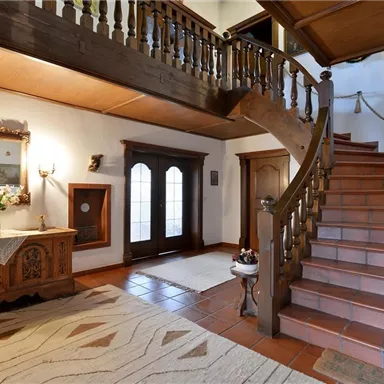 Ein einladender Eingangsbereich mit einer geschwungenen Treppe und Holzverkleidung. Der Raum ist gemütlich mit einem schönen Teppich und einer Blumenarrangement dekoriert.