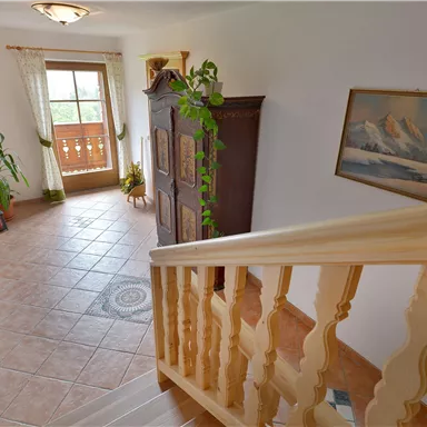 Ein freundlicher Flur mit einer Holztreppe und einem großen Fenster. Pflanzen und ein Gemälde verleihen dem Raum eine einladende Atmosphäre.