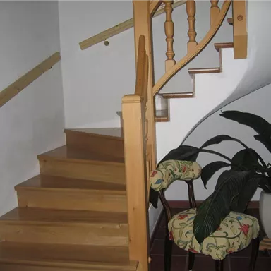 A wooden staircase with a yellowish handrail leads upstairs. Next to the stairs stands a chair with a colorful cushion and a plant in a pot.