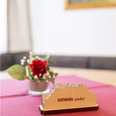 Ein Tisch mit einer roten Tischdecke, auf dem sich eine kleine Blumenvase mit einer Rose und einer dekorativen Holzfigur mit der Aufschrift "GOING stärkt!" befindet. Im Hintergrund ist ein verschwommenes Bild zu sehen.