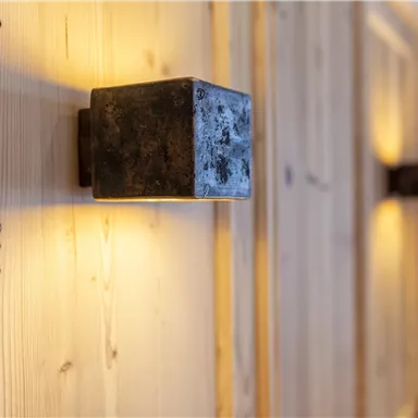 Eine moderne Wandleuchte aus Metall strahlt ein warmes Licht aus. Der Hintergrund besteht aus hellem Holz, was eine gemütliche Atmosphäre schafft.