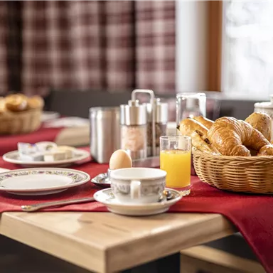 Ein gemütlicher Frühstückstisch mit frischen Croissants, einer Tasse Kaffee und einem Glas Orangensaft. Der Tisch ist mit einer roten Tischdecke gedeckt und eine freundliche Atmosphäre strahlt aus.