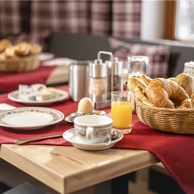 Ein schön gedeckter Frühstückstisch mit einem Korb voller frischer Croissants. Dazu Tassen, Gläser und ein rotes Tischdecke für eine gemütliche Atmosphäre.