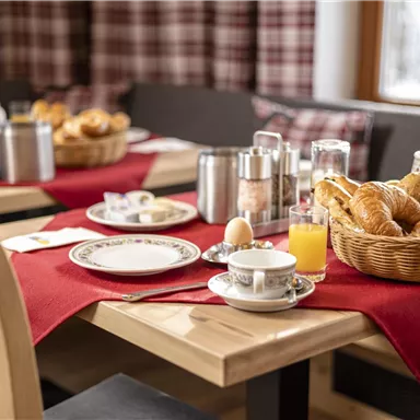 Ein gemütlicher Frühstückstisch mit frischen Brötchen, Croissants und einem Glas Orangensaft. Die Tische sind mit roten Tischdecken dekoriert und es gibt eine Auswahl an Geschirr und Getränken.