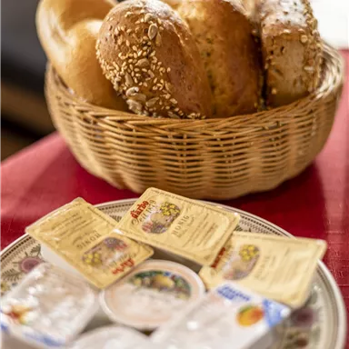 Ein Korb mit verschiedenen Brötchen steht auf einem Tisch. Daneben sind mehrere kleine Portionen Butter und Marmelade auf einem Teller arrangiert.