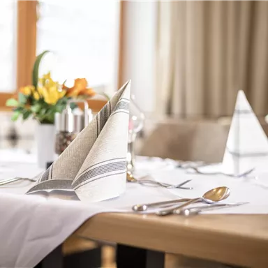 Ein schön gedeckter Tisch mit eleganten Servietten und Besteck. Im Hintergrund sind frische Blumenarrangements zu sehen.