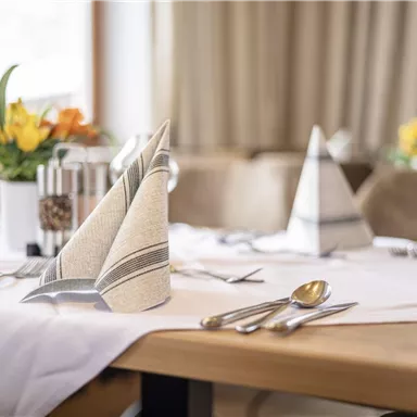 Ein elegant gedeckter Tisch mit servierten Bestecken und kunstvoll gefalteten Servietten. Frische Blumen in der Mitte des Tisches schaffen eine einladende Atmosphäre.