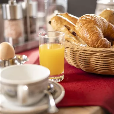 Ein gedeckter Frühstückstisch mit Croissants, einem gekochten Ei und einem Glas Orangensaft. Im Hintergrund sind Salz- und Zuckergefäße zu sehen.