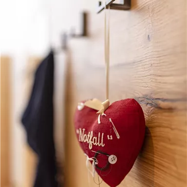 Ein rotes Stoffherz mit der Aufschrift „Notfall“ hängt an einem Haken an einer Holzwand. Daneben hängen dunkle Kleidungsstücke.