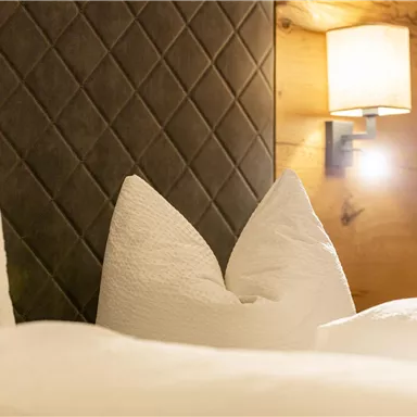 Ein gemütliches Bett mit großen, weißen Kissen und einem modernen Nachttischlicht. Die Wand ist aus Holz und hat ein elegantes, diagonales Muster.