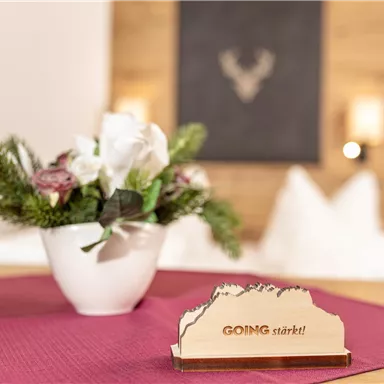 Ein schöner Tisch mit einer blühenden Pflanze in einem weißen Topf und einer dekorativen Holzauflage mit der Aufschrift "GOING startet!". Im Hintergrund ist eine moderne, gemütliche Atmosphäre zu sehen.