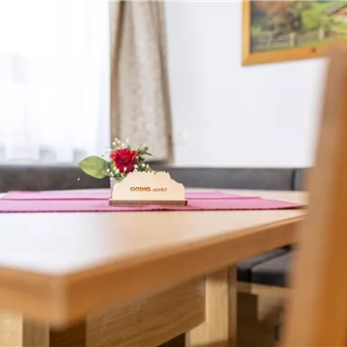 Ein Holzstuhl und ein Tisch mit einer roten Tischdecke. Auf dem Tisch steht eine kleine Blumendekoration mit einer Karte.