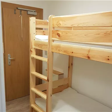 A simple room with a bunk bed made of light wood. The wall is neutral, and a door leads outside.