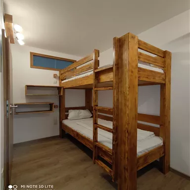 Ein Zimmer mit einem stabilen Etagenbett aus Holz. Die Wände sind hell, und es gibt ein großes Fenster sowie eine kleine Ablage.