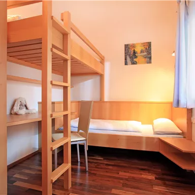 Ein schlichtes Zimmer mit einem Hochbett und einem Einzelbett. Die Wände sind hell und das Licht strömt durch das Fenster.