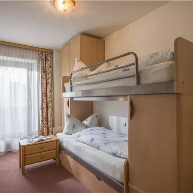 A bright room with a bunk bed and a single bed. The windows are decorated with blooming curtains, and the room feels inviting and cozy.