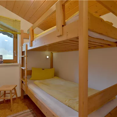 Ein einfaches Schlafzimmer mit einem Etagenbett aus Holz. Der Raum hat ein Fenster, einen Hocker und ist hell und freundlich gestaltet.