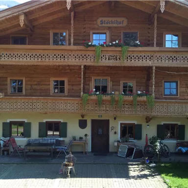 Ein traditionelles Holzhaus mit Balkon und blühenden Pflanzen. Es steht in einer ländlichen Umgebung mit grünen Bäumen und einem klaren Himmel.