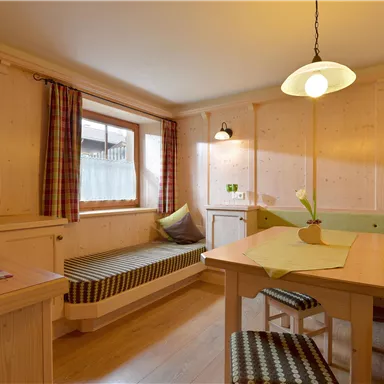 Eine gemütliche Wohnstube mit Holzvertäfelung, einem Tisch und Sitzgelegenheiten. Ein Fenster sorgt für Tageslicht und eine Uhr ist an der Wand angebracht.