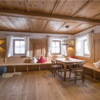 A cozy room with wooden walls and a light wood floor. In the center, there is a table with chairs and benches, decorated with red cushions.