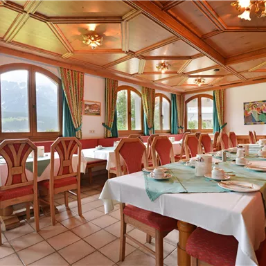 Ein einladender Essbereich mit Tischen und Stühlen in einem rustikalen Stil. Große Fenster bieten einen Blick auf die Berge und farbenfrohe Wandbilder ergänzen die Einrichtung.