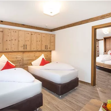 A cozy room with two single beds and traditional wooden design. The room is bright and inviting, with an open view of an adjacent room.