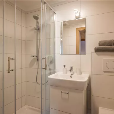 Ein modernes Badezimmer mit einer Dusche aus Glas und einem Waschbecken. Frische Handtücher und Pflegeprodukte sind auf der gesamten Fläche arrangiert.