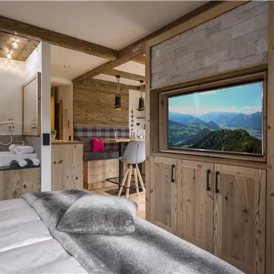 A modern room with wooden furniture and a cozy bed. In the background, a television displaying a landscape can be seen.