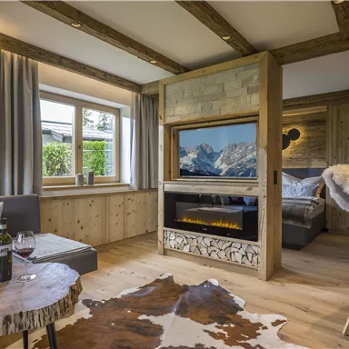 A cozy living space with wooden details and a modern fireplace. There is a seating area with wine and a view of the mountains.