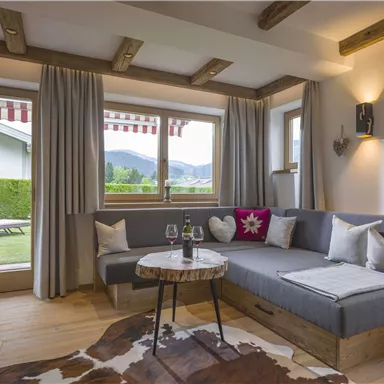 A cozy living area with a large sofa corner and a coffee table. Large windows offer a view of the garden and the mountains.